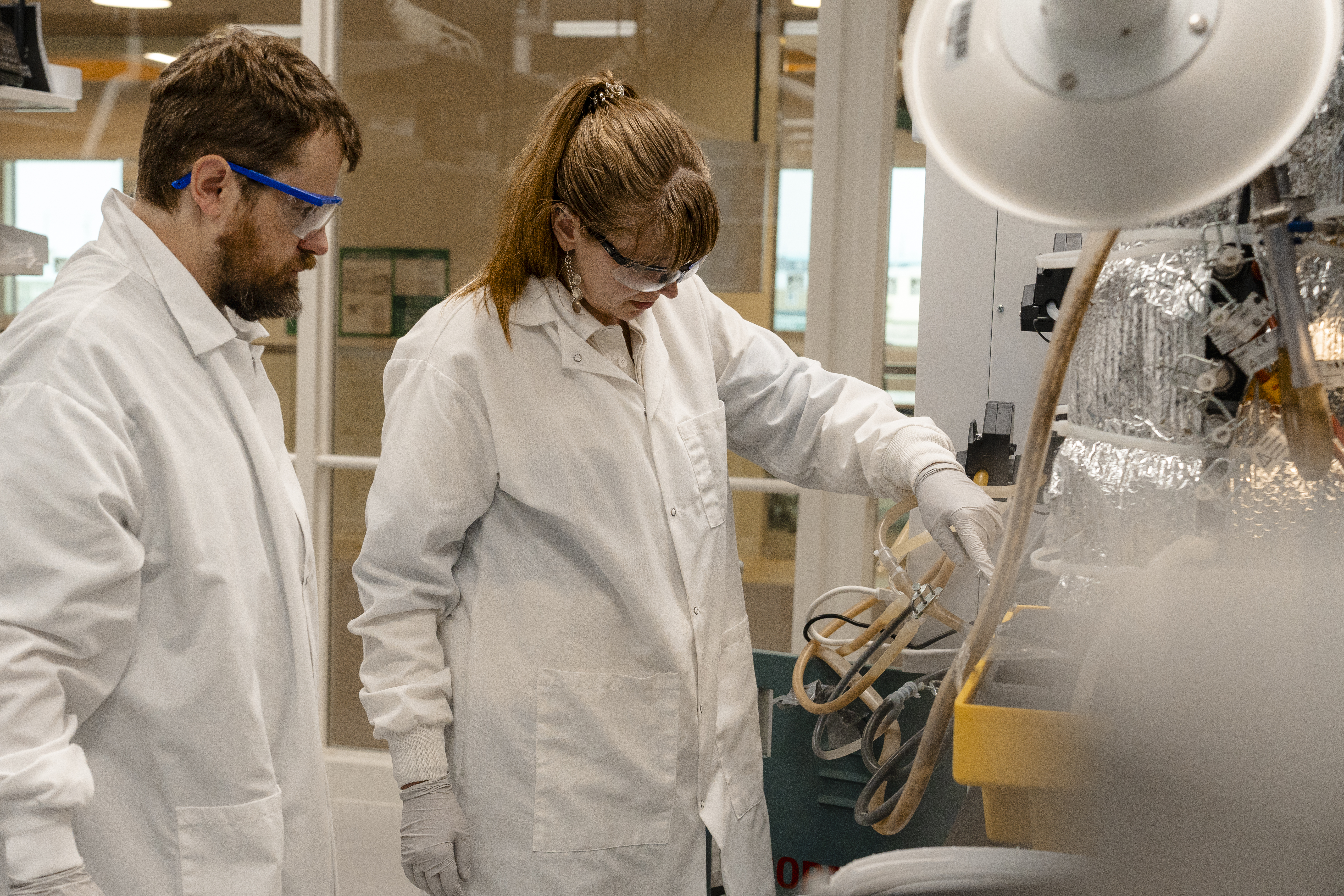 A man in a lab coat watching a woman in a lab coat pointing to lab equipment.